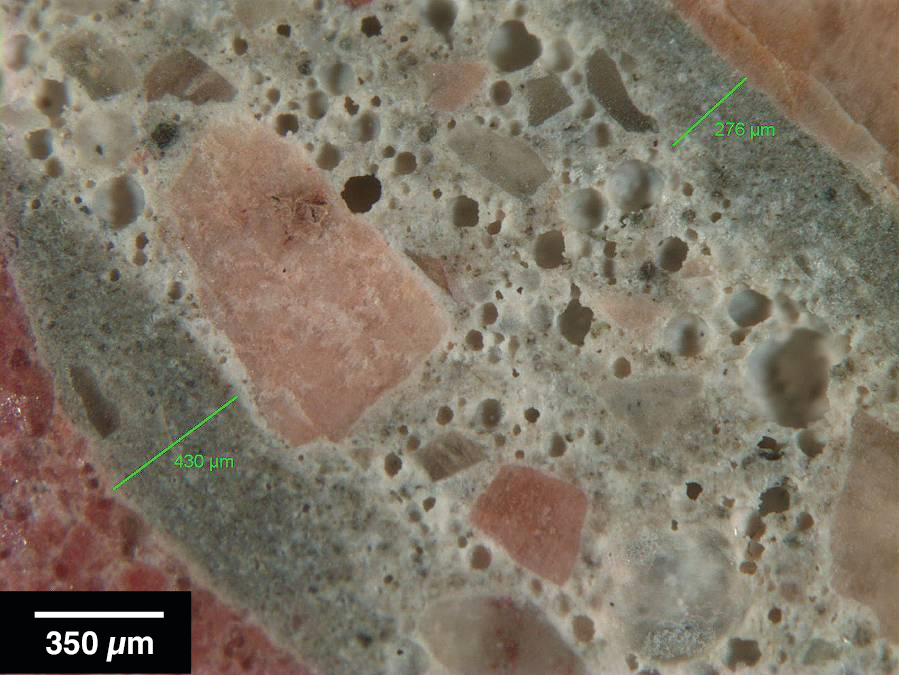 Figure 1: Reflected light photomicrograph of polished surface showing dark, low w/cm mortars (green bars) coating aggregate particles. These are typical indicators of retempering.