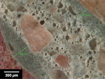 Figure 1: Reflected light photomicrograph of polished surface showing dark, low w/cm mortars (green bars) coating aggregate particles. These are typical indicators of retempering.