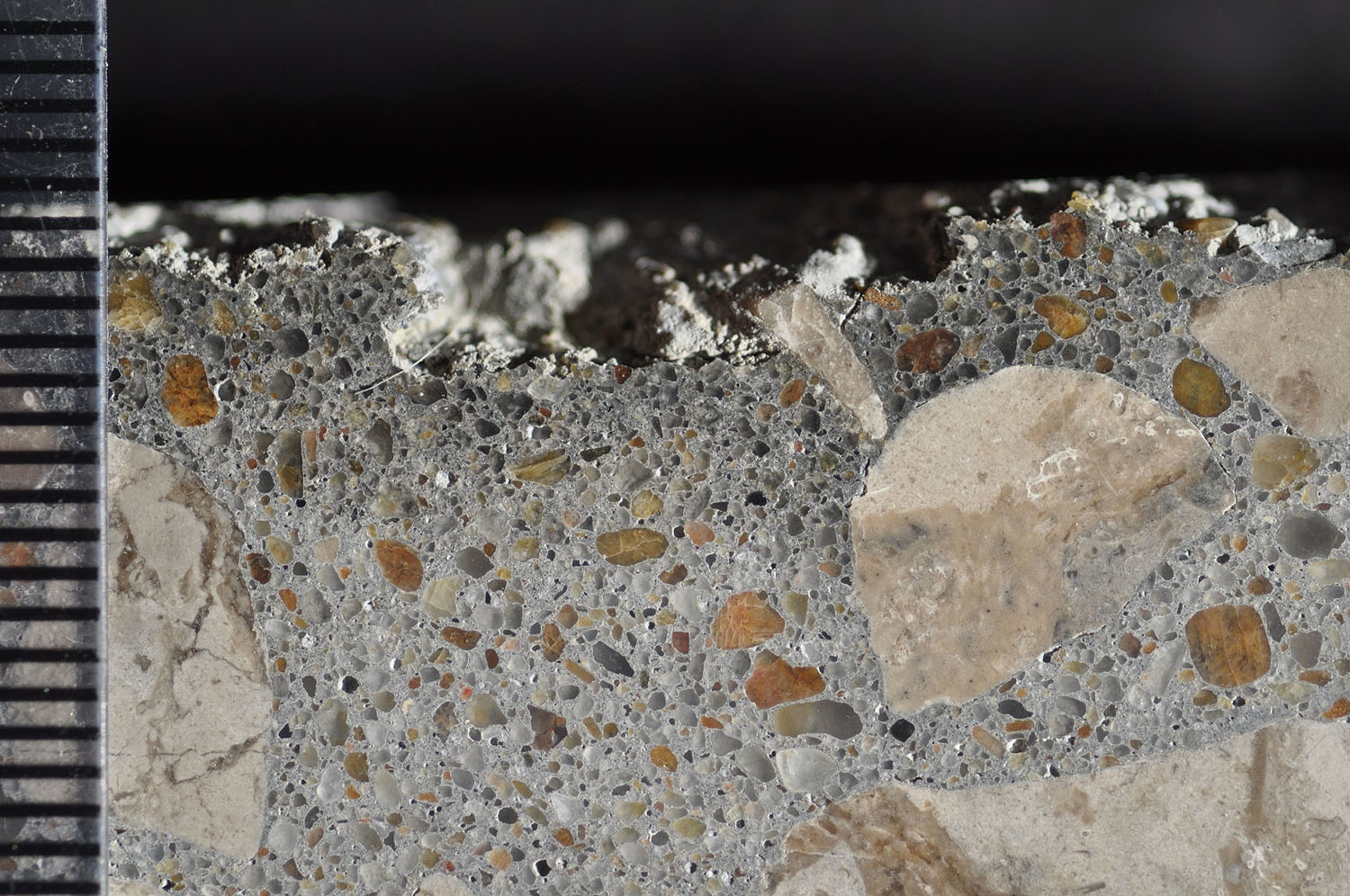 Photograph of polished surface from a concrete core removed from a poorly cured slab on ground showing layer of dark paste at the top of the core. Scale in millimeters. The loss of material at the top is due to poor hydration.