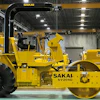 Yellow Sakai industrial road roller in manufacturing facility with overhead crane