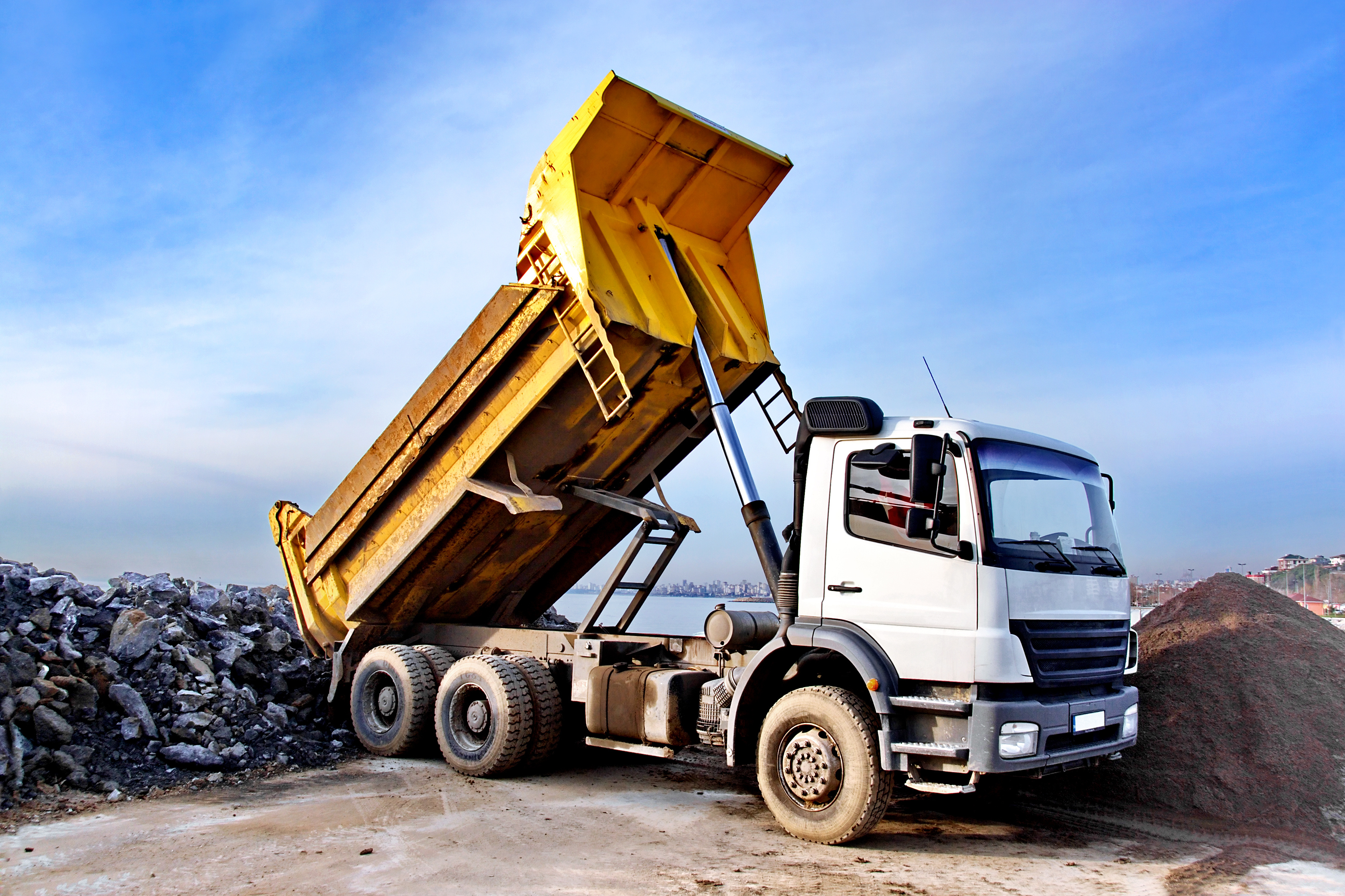 Yellow-topped dump truck unloading rocks at construction site with city skyline