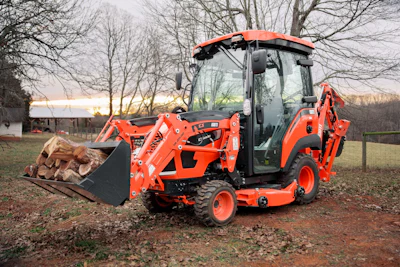 KIOTI CS30 sub-compact tractor with front loader carrying firewood logs in rural setting