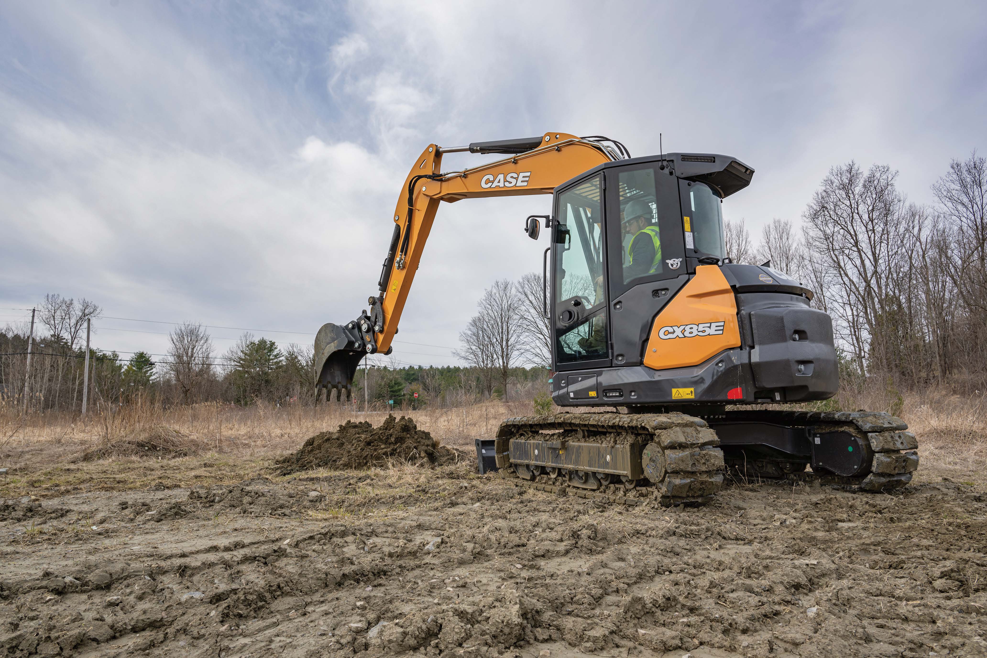 Case excavator digging in muddy field with bare trees and overcast sky