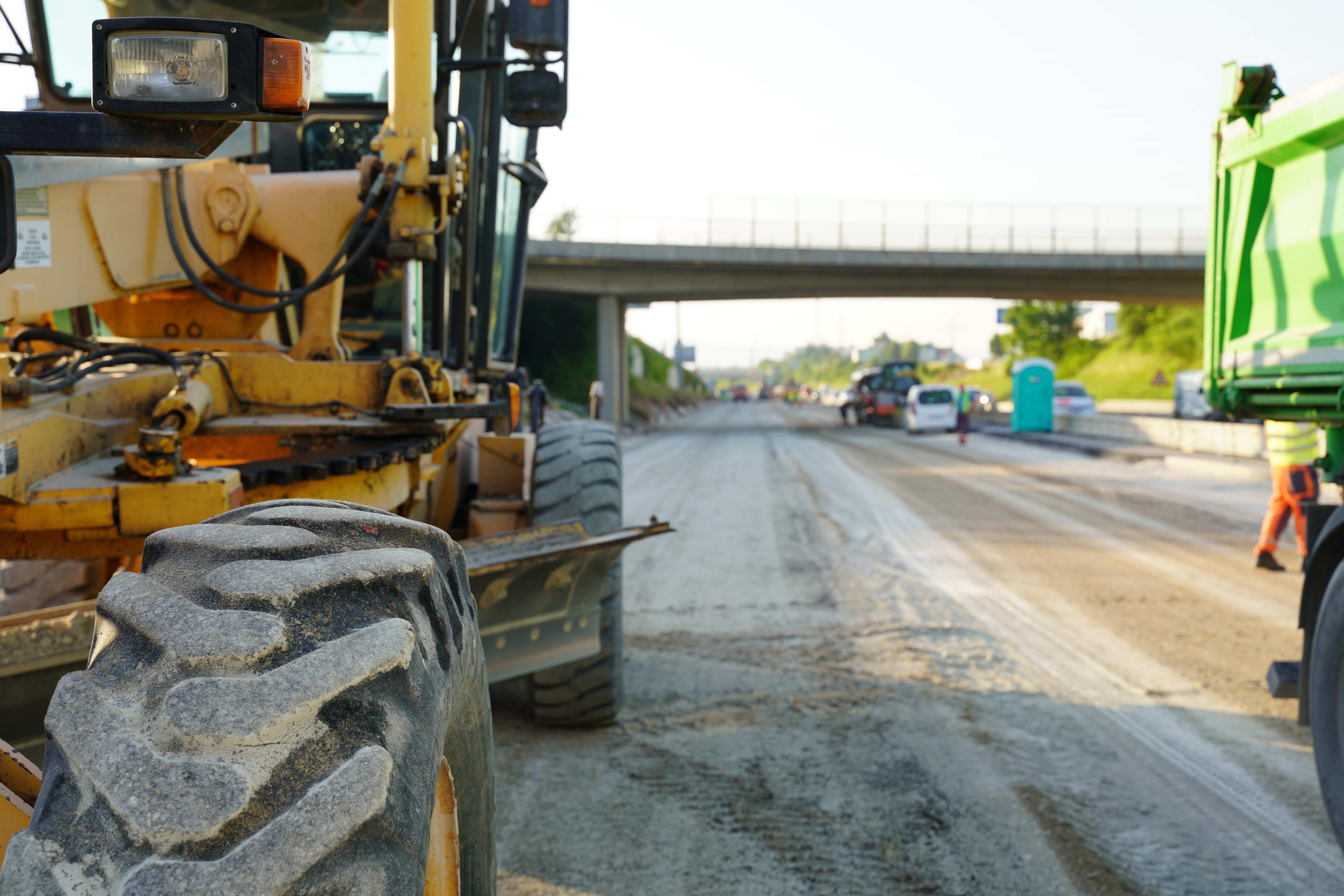 Yellow motor grader on road construction site with highway overpass