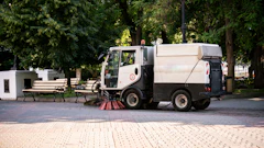 Street sweeping truck cleaning urban park pathway with trees and benches