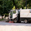 Street sweeping truck cleaning urban park pathway with trees and benches