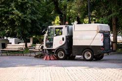 Street sweeping truck cleaning urban park pathway with trees and benches