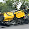 Yellow Bomag road paving machine spreading fresh asphalt on street with workers