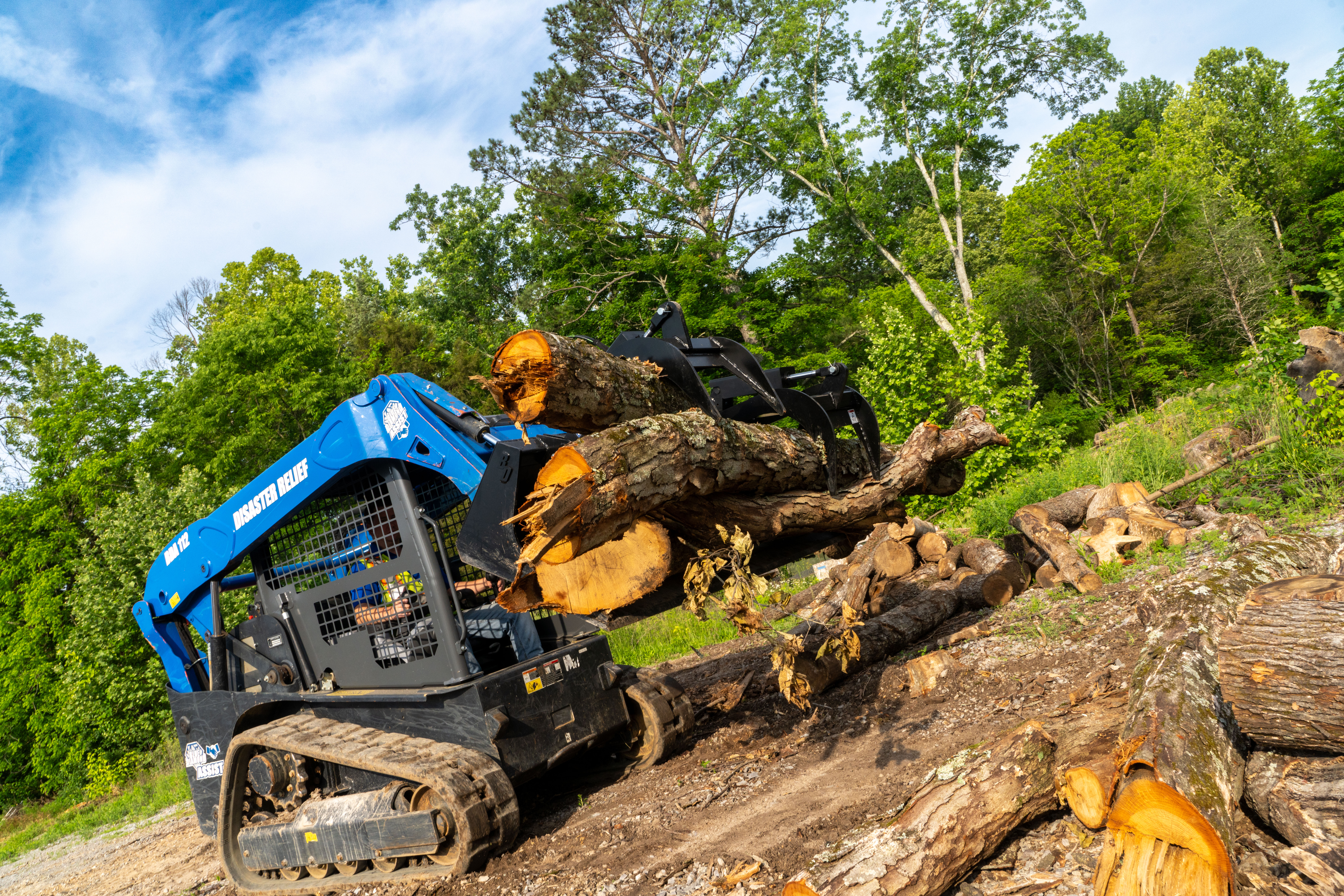 Blue tracked skid steer loader moving large fallen tree logs on rocky terrain with green forest background
