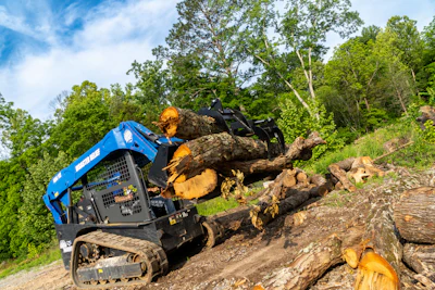 Blue tracked skid steer loader moving large fallen tree logs on rocky terrain with green forest background