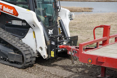 White Bobcat skid-steer loader with red trailer on muddy ground near water