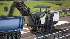 Road construction machine milling asphalt on highway with blue dump truck