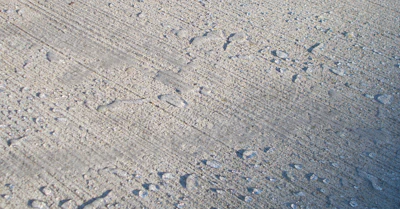 Textured concrete or sandy surface with scattered small rocks and linear markings