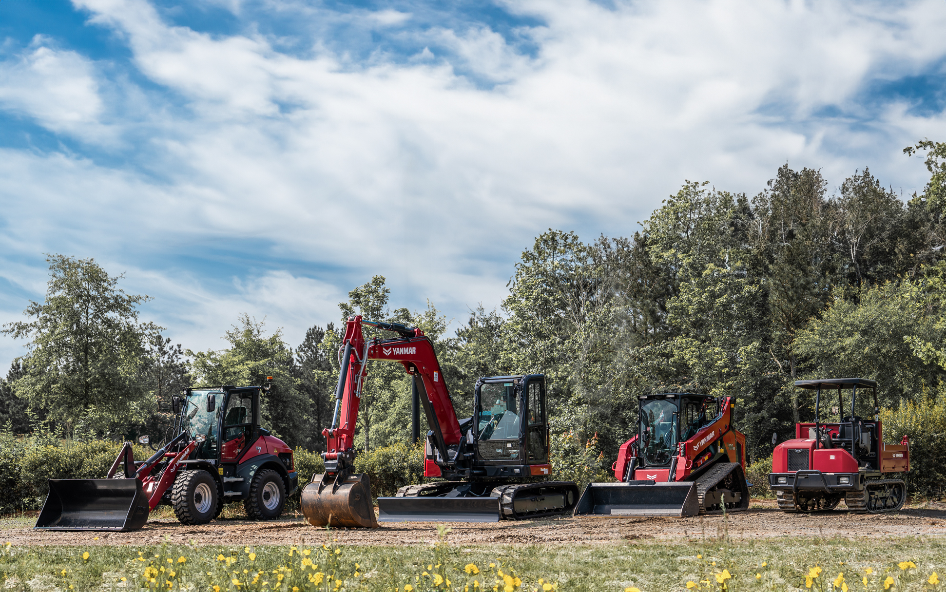 Red and gray construction vehicles lined up in grassy field with trees