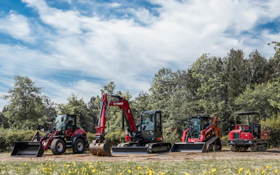 Red and gray construction vehicles lined up in grassy field with trees