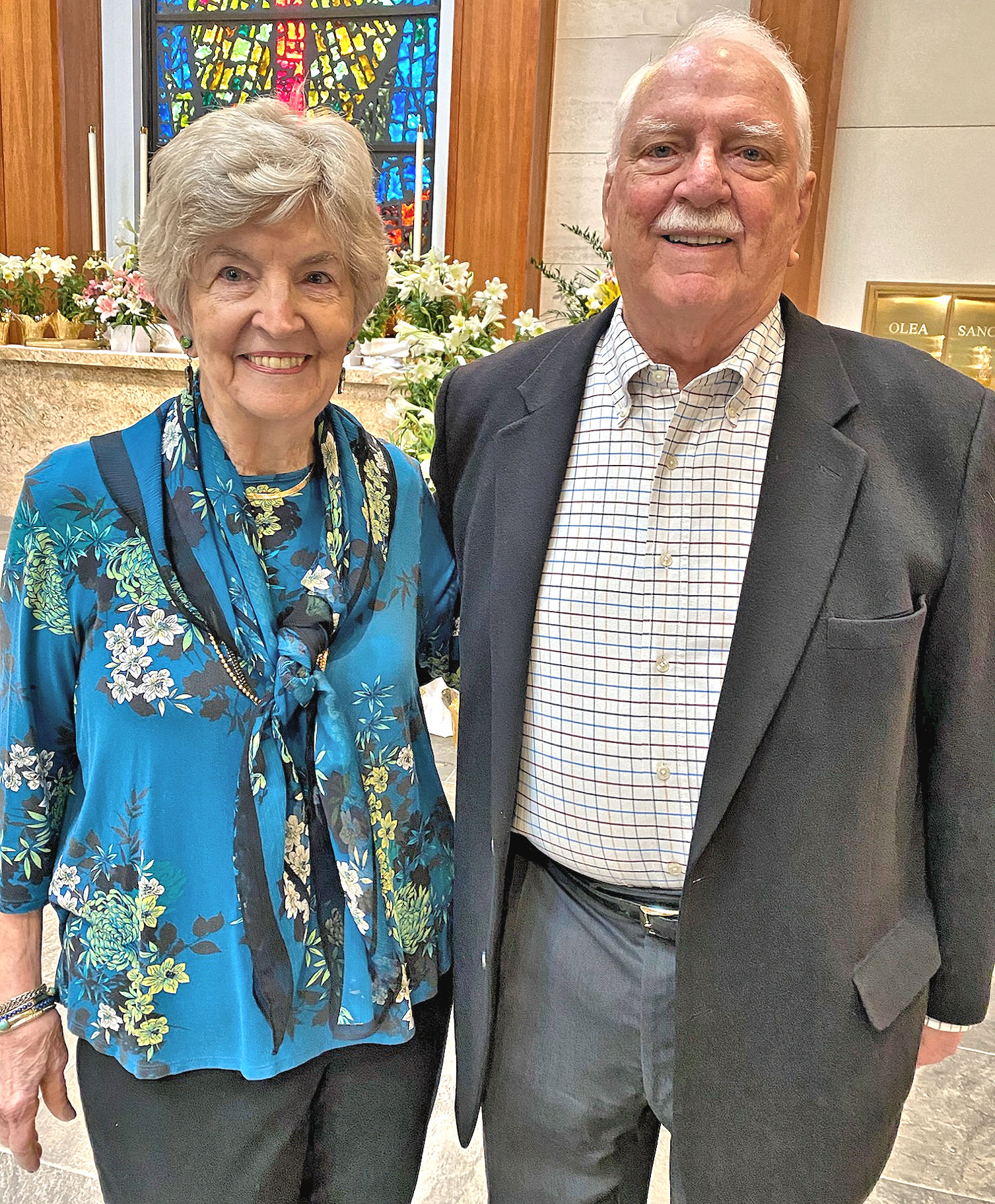 Smiling senior couple dressed formally, standing together in church setting with flowers