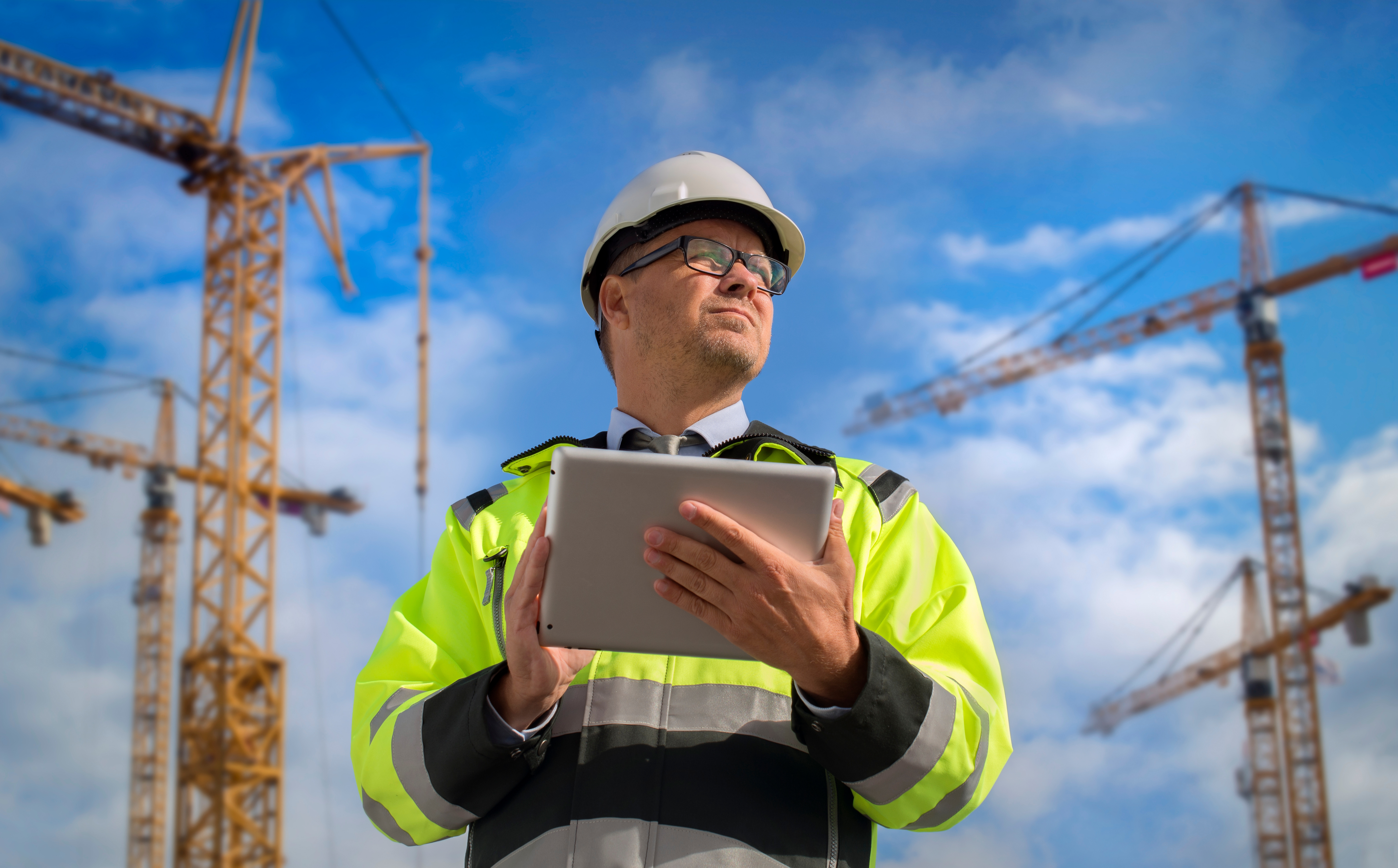 Construction manager in safety vest and hard hat using tablet at crane-filled worksite