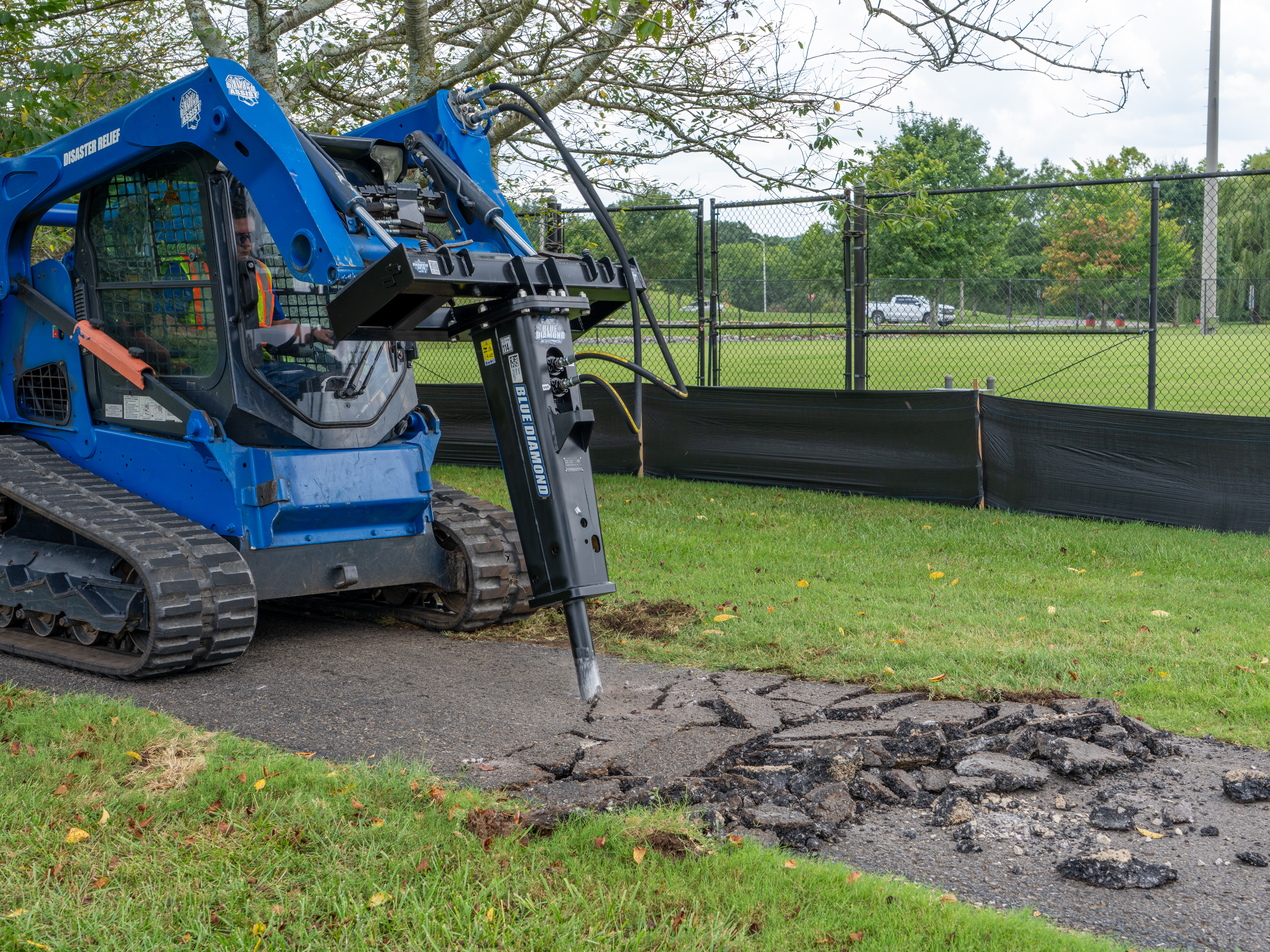 Blue tracked construction vehicle breaking concrete pavement near grassy field