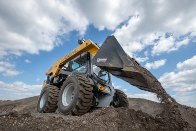 Yellow skid steer loader dumping dirt on rocky terrain with cloudy sky