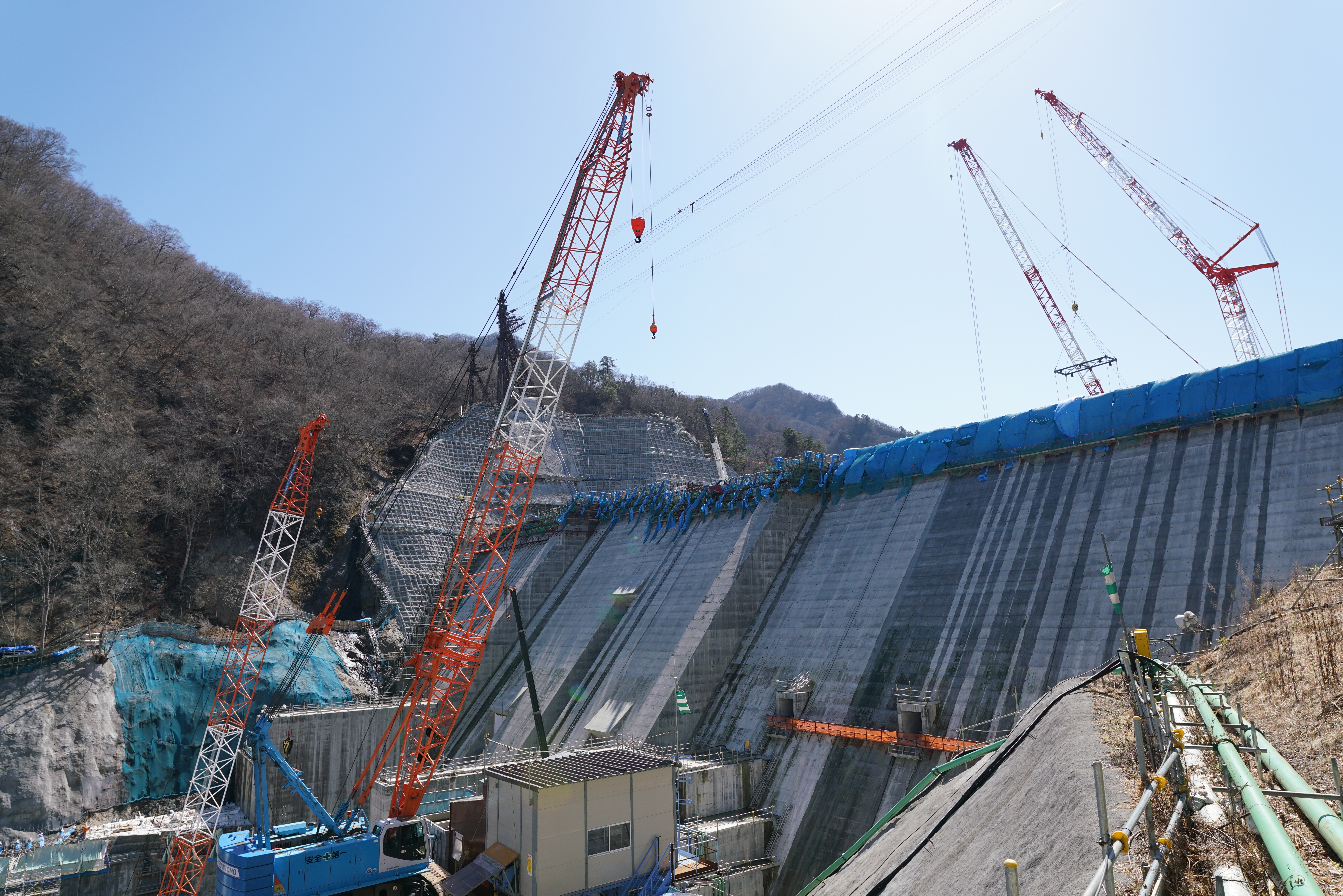 Large concrete dam under construction with red and white cranes on mountainside