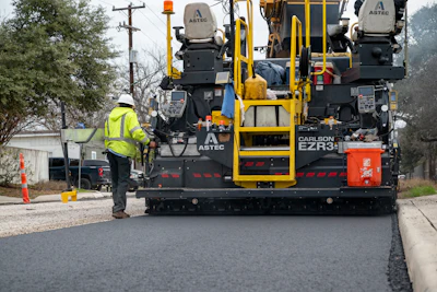 Road paving machine laying fresh asphalt on urban street with worker in safety vest