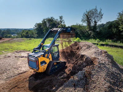 Yellow Hyundai tracked skid steer loader moving dirt on a construction site with green field background