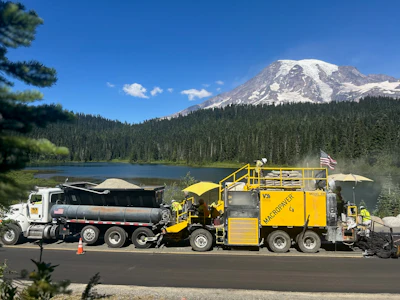 Yellow road paving machine and truck near lake with snow-capped Mount Rainier in background