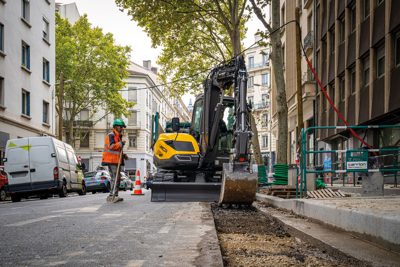 Yellow excavator digging street with construction worker in orange safety vest
