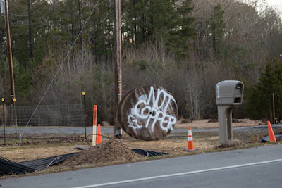 Construction site with traffic cones, utility pole, and graffitied metal cylinder near wooded area
