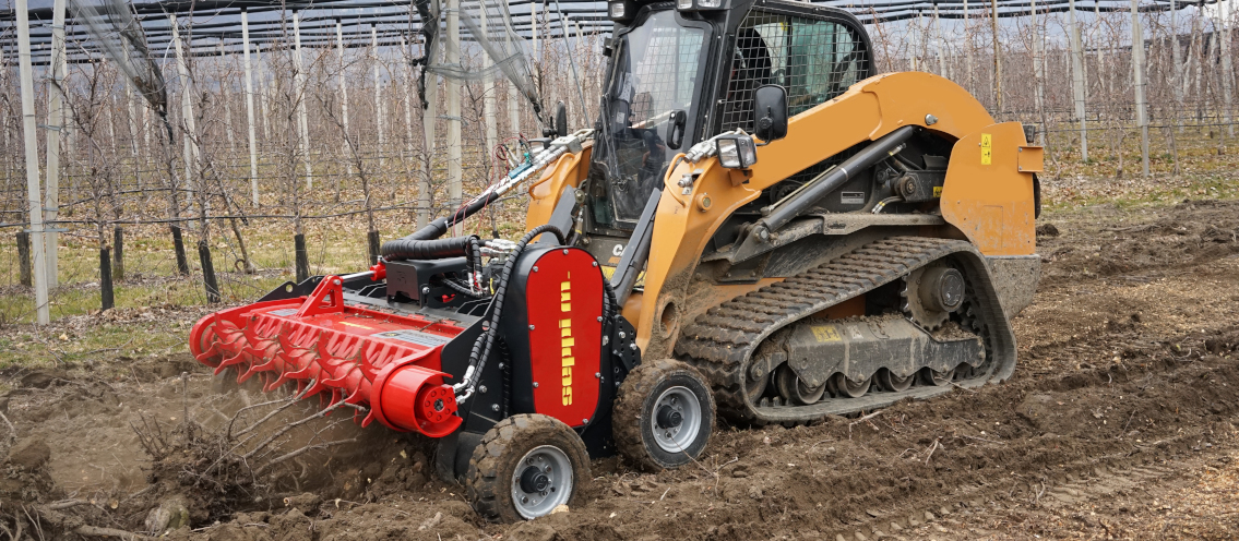 Yellow tracked skid steer with red forestry mulcher working in bare orchard