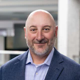 Smiling professional in navy blazer and checked shirt, bald with gray beard