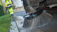 Street sweeper truck cleaning curb with large rotating brush, worker in high-visibility vest nearby