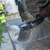 Street sweeper truck cleaning curb with large rotating brush, worker in high-visibility vest nearby