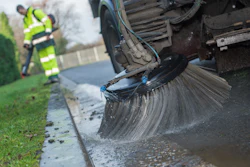 Street sweeper truck cleaning curb with large rotating brush, worker in high-visibility vest nearby
