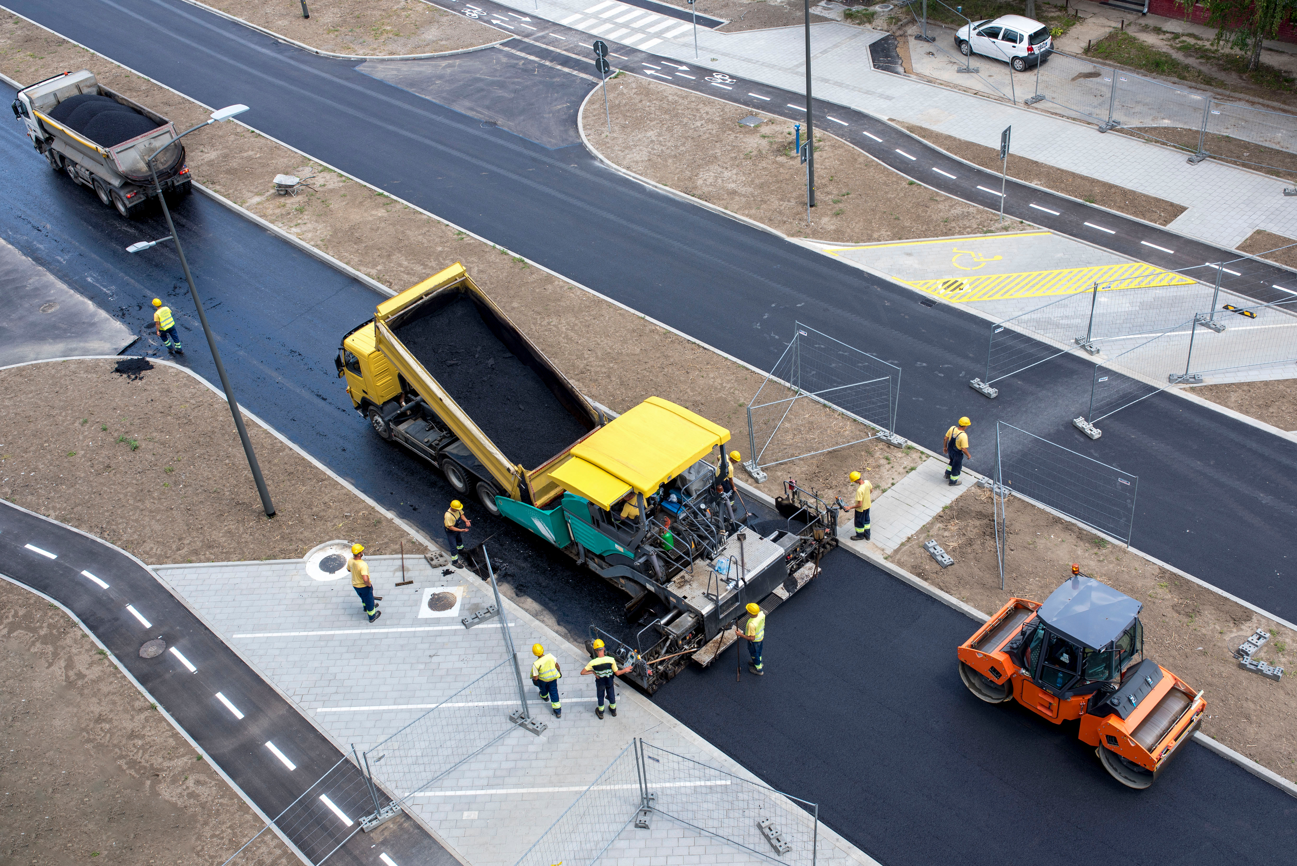 Road construction site with yellow paver, orange roller, and workers laying fresh asphalt