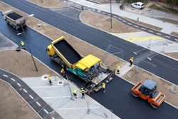 Road construction site with yellow paver, orange roller, and workers laying fresh asphalt