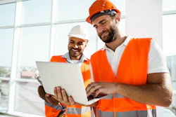 Two construction workers in safety vests and hard hats collaborate on a laptop