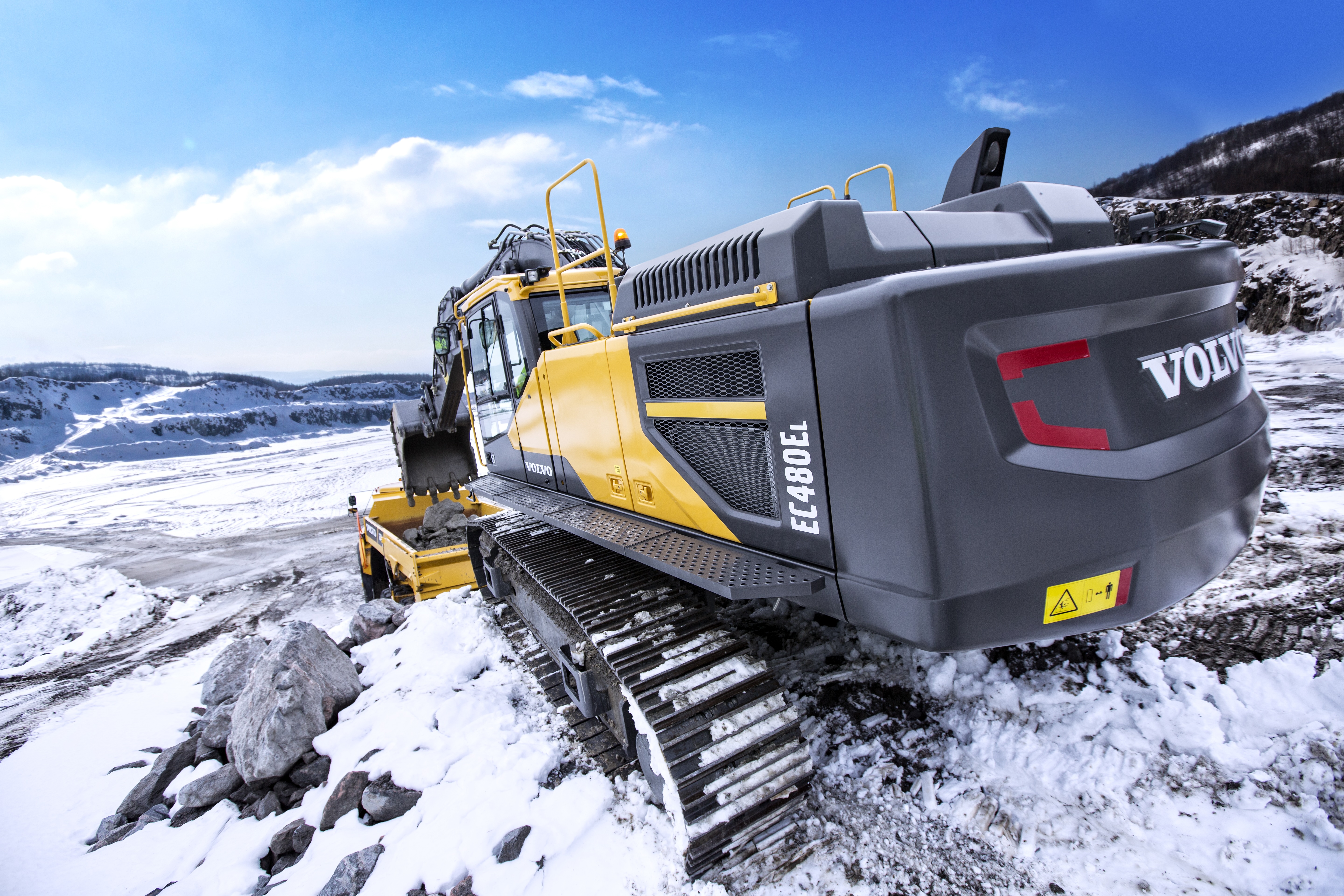 Yellow Volvo excavator on rocky snow-covered terrain with mountain backdrop