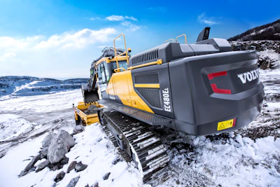 Yellow Volvo excavator on rocky snow-covered terrain with mountain backdrop