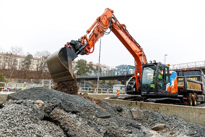 Orange excavator moving gravel at urban construction site with buildings in background
