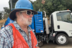 Construction worker in blue hard hat and orange safety vest near street sweeping truck