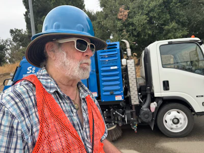 Construction worker in blue hard hat and orange safety vest near street sweeping truck