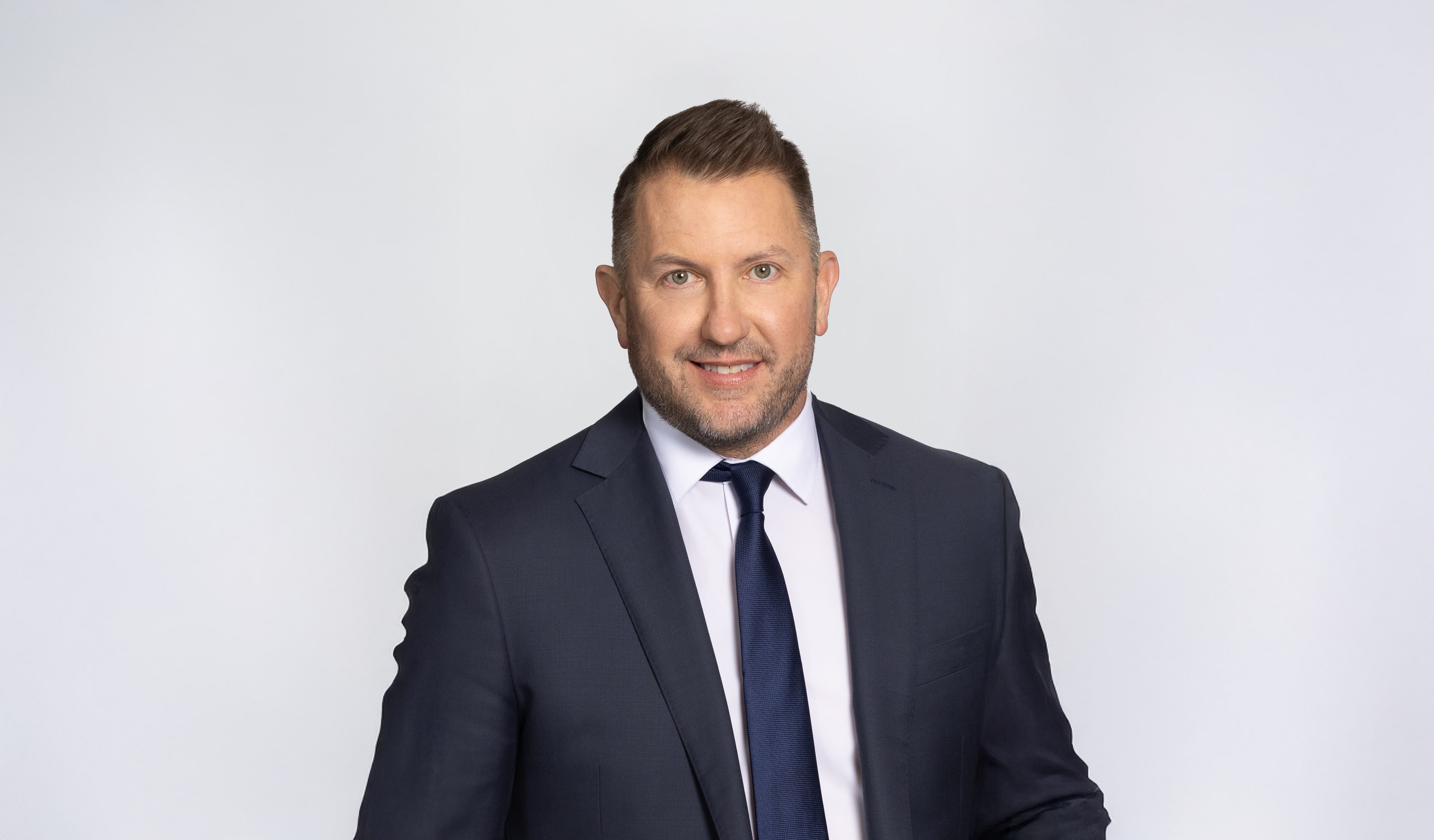 Professional man in dark suit and navy tie smiling against neutral background