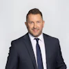 Professional man in dark suit and navy tie smiling against neutral background