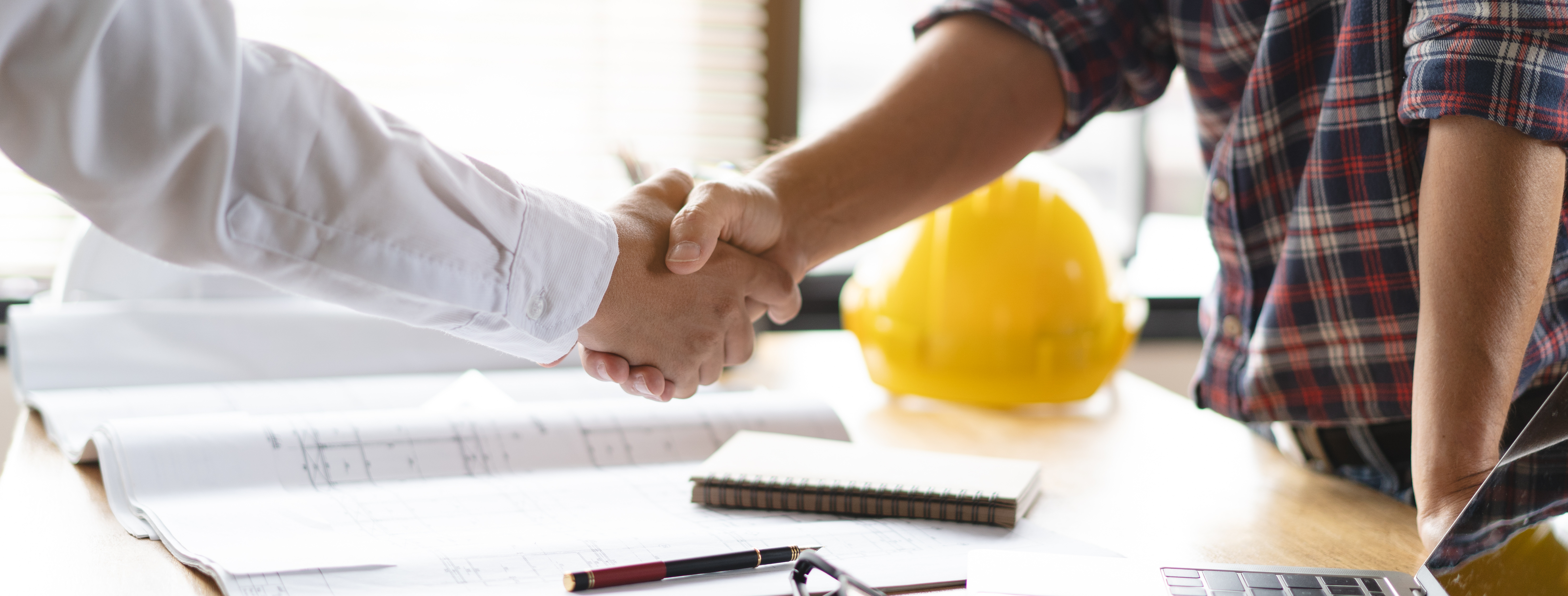 Construction professionals shaking hands over work documents with yellow hard hat