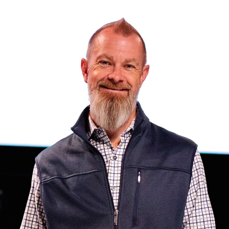 Middle-aged man with gray beard smiling, wearing navy vest and plaid shirt Headshot