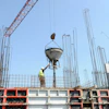 Construction worker guides concrete mixer suspended by red crane at building site with steel reinforcement framework