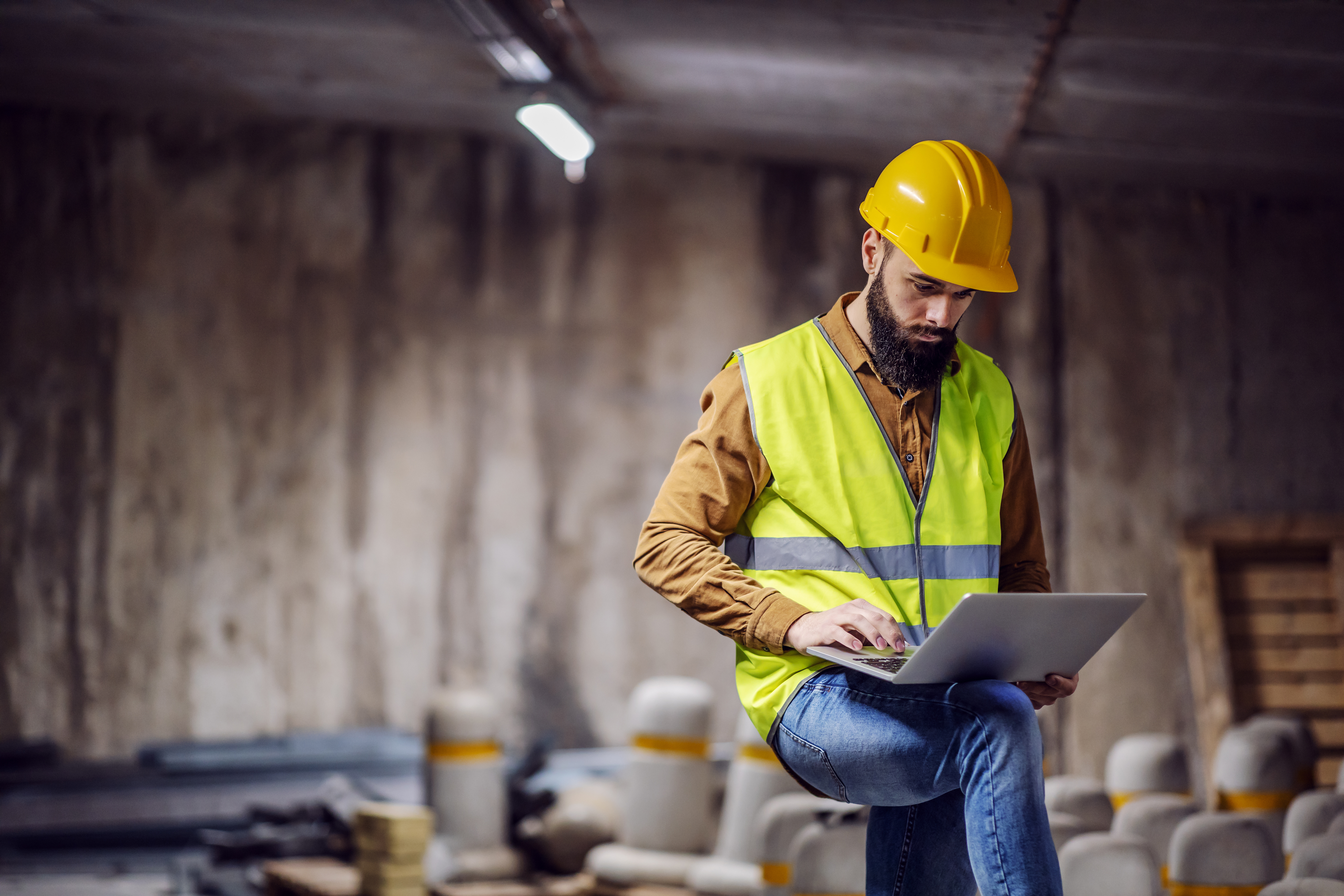Construction worker in yellow hard hat and safety vest working on laptop at industrial site