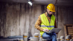 Construction worker in yellow hard hat and safety vest working on laptop at industrial site