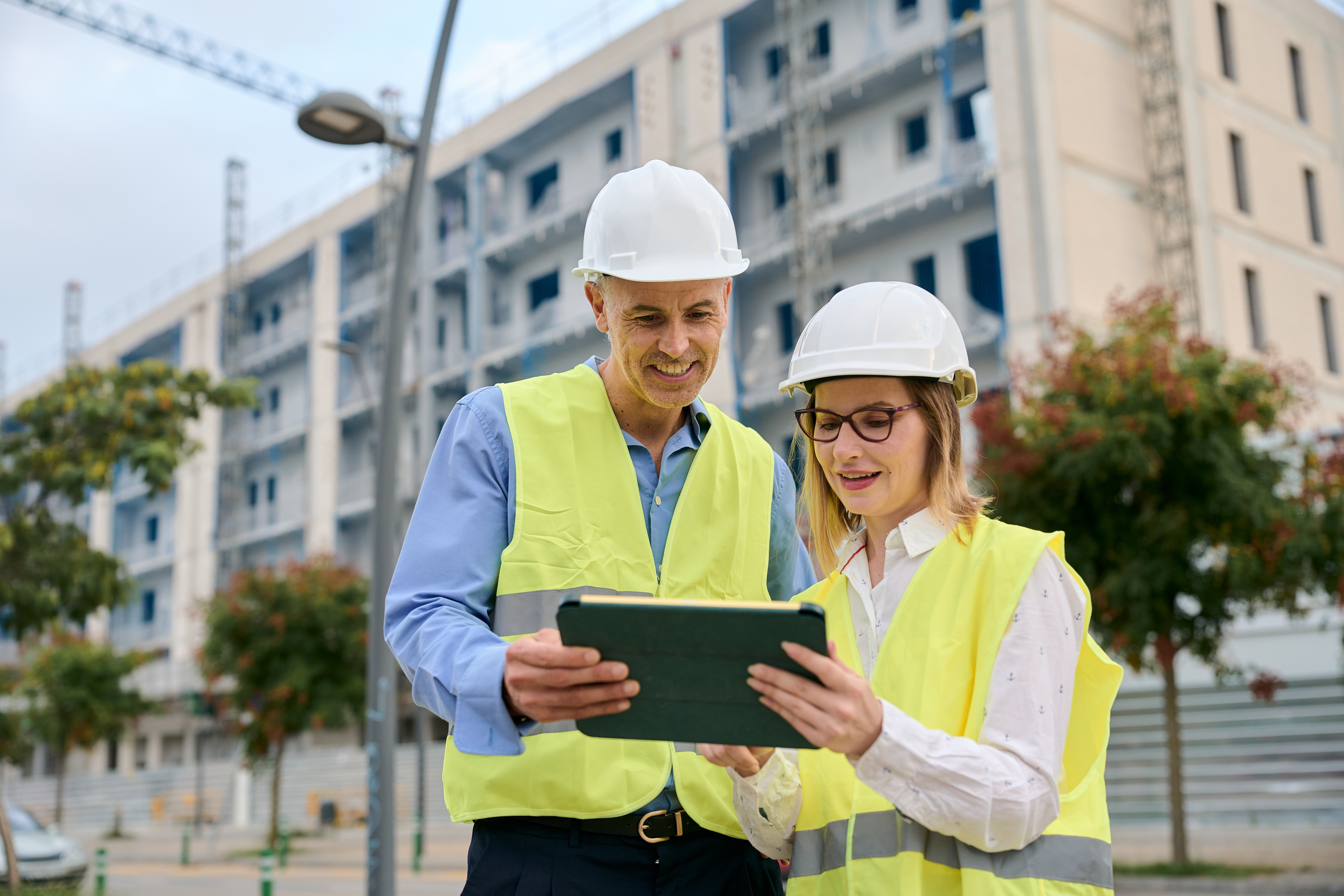Construction workers in safety vests and hard hats reviewing tablet near apartment buildings